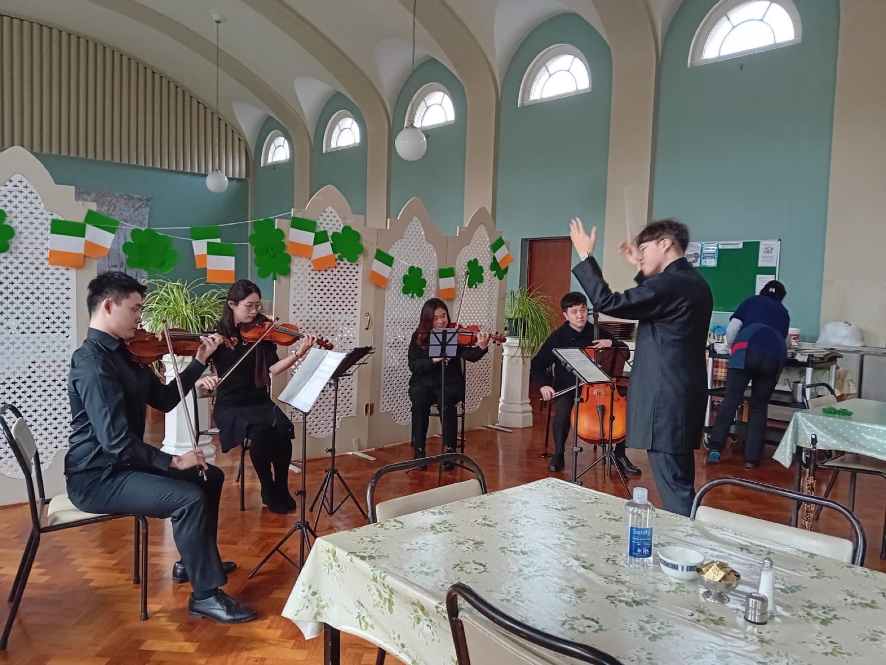 Four string players, seated and playing, and their conductor in a hall hung with Irish flags and shamrocks
