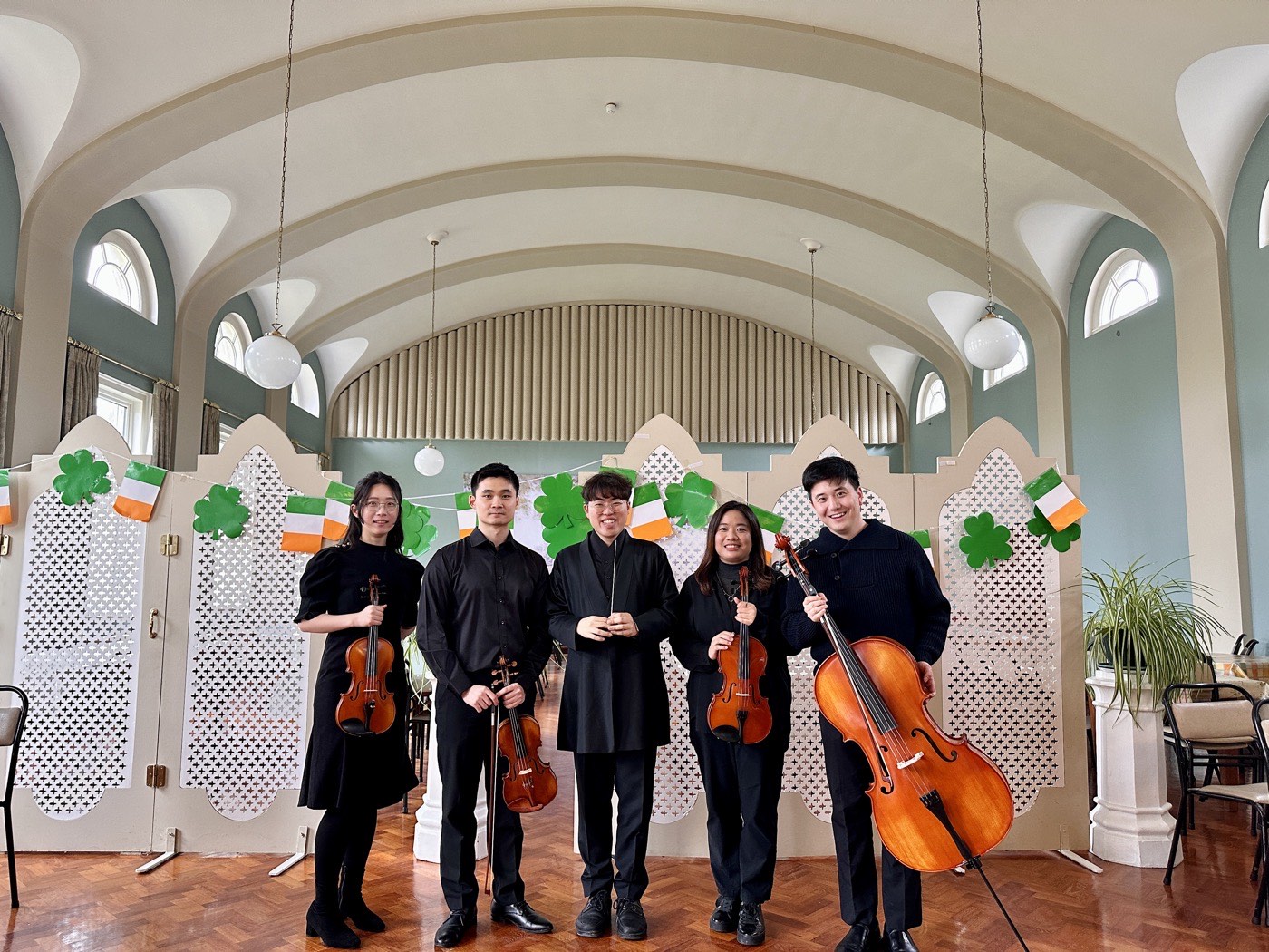 Four string players and their conductor standing with their instruments in a hall hung with Irish flags and shamrocks