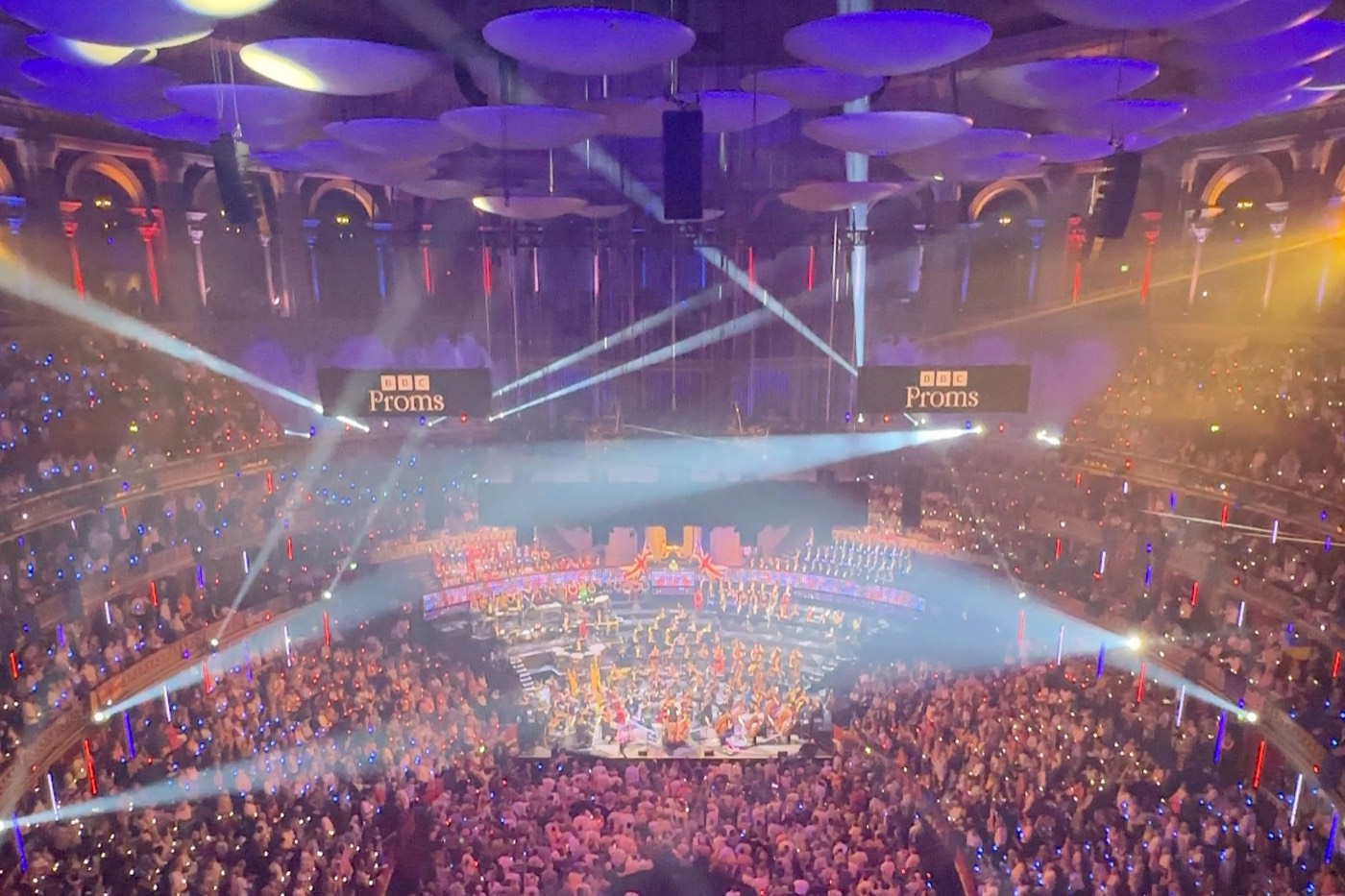 A full Royal Albert Hall during a BBC Prom, seen from high in the audience: the orchestra on a lit stage, beams of light and the acoustic discs overhead
